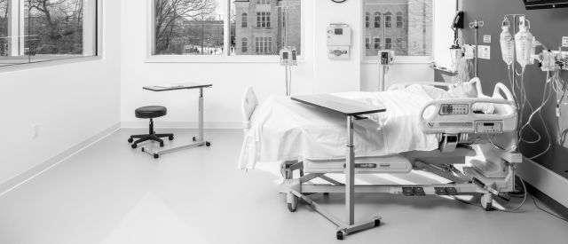 一个现代人的单色照片, 空的医院房间里有一张床, various medical equipment, 还有一扇可以俯瞰外面建筑的大窗户.